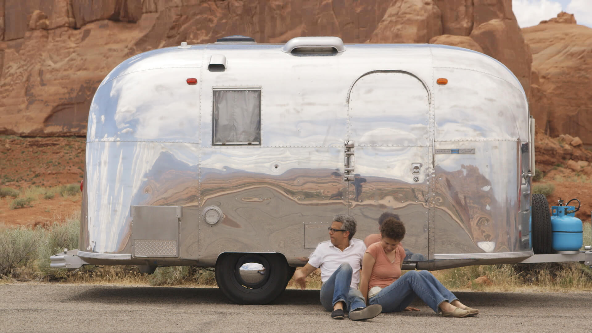 Still from Unrest. A man and a woman sitting on the ground in front of a small, silver airstream trailer. The background is the mountains of a red, sandy desert.