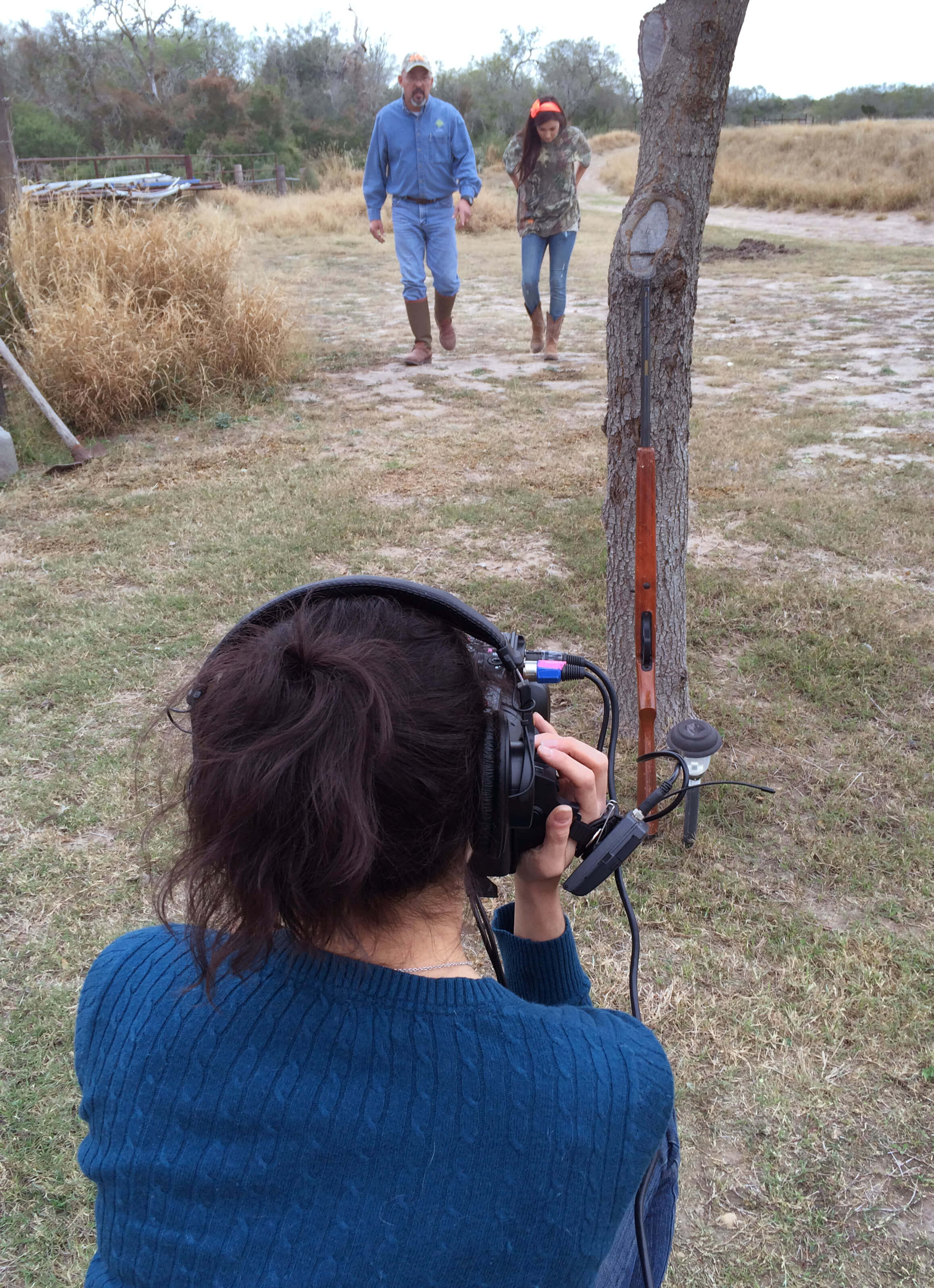 Production still from Hot Girls Wanted. A woman is seen from behind, and she is wearing headphones and holding a camera that is pointed in front of her towards a man and an adolescent girl walking side by side.