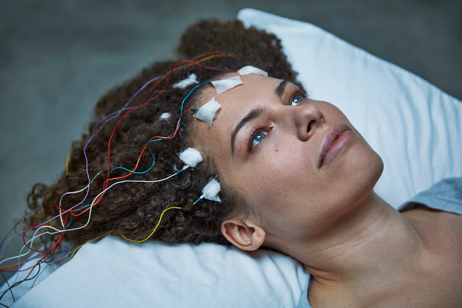 Still from Unrest. A woman's head is lying on a pillow with sensors taped to her forehead, the wires laying on her hair.