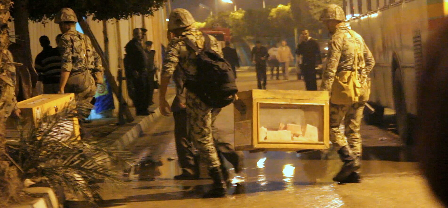 Still from The Vote. People in the street watch as soldiers carry boxes with votes inside of them.