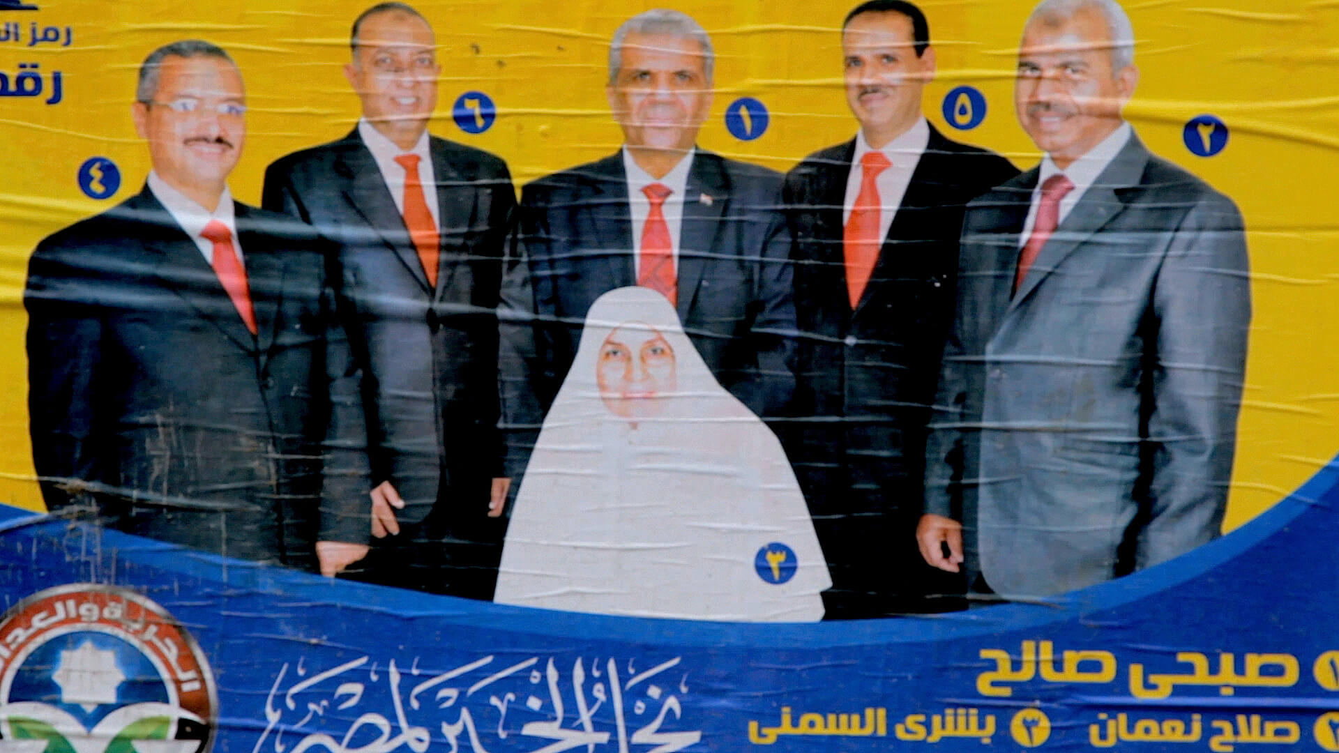 Still from The Vote. Photograph of a poster pasted on a wall. Three persons in suites are behind a person wearing a hijab.