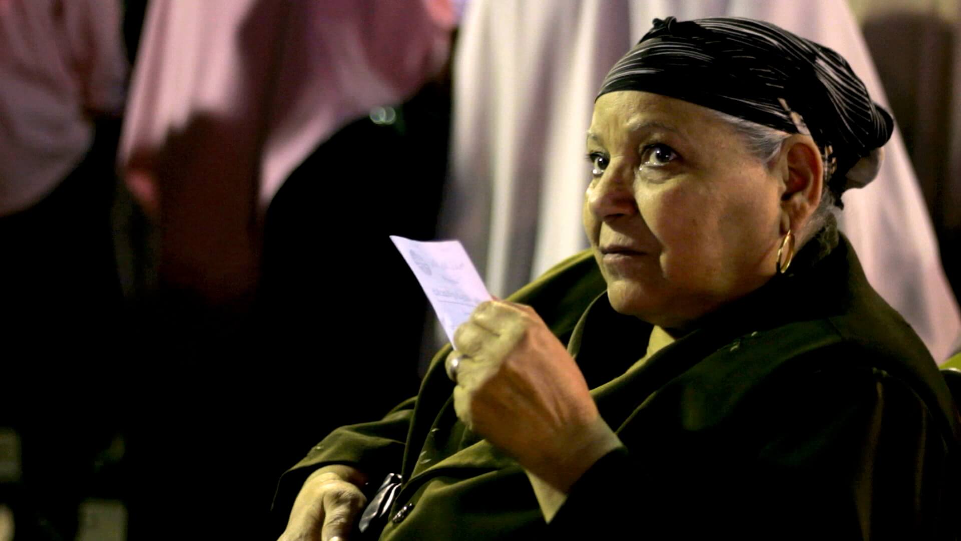 Still from The Vote. A woman wearing a scarf on her head holds a piece of paper in her hands. She looks up from the camera.