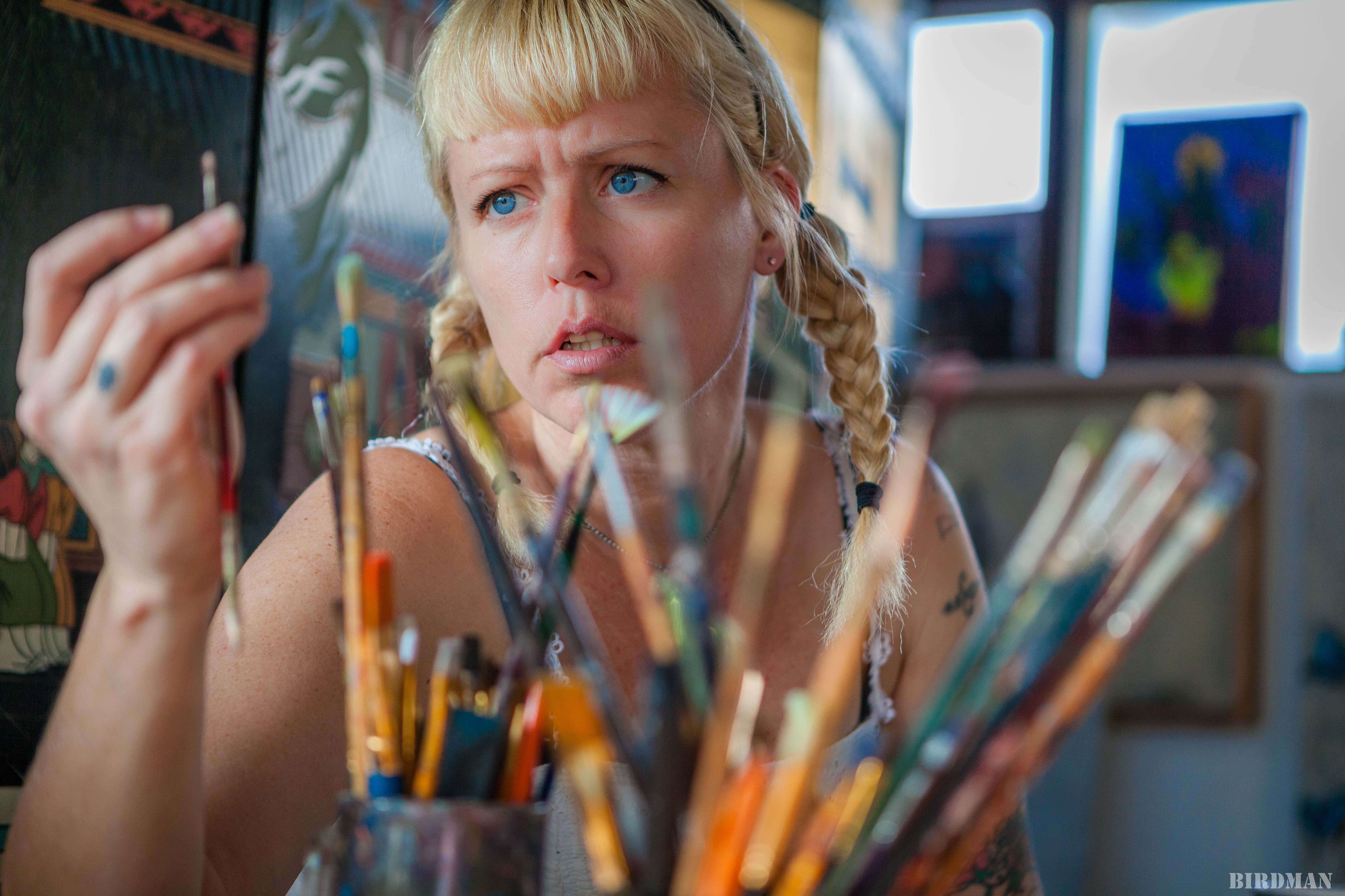 Lydia Emily from the chest up sitting behind many paintbrushes in cups, staring with furrowed brows at a brush in her hand.