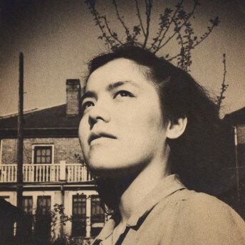 Archival photo in a sepia tone of young Grace Lee Boggs. The photograph is taken in a low-angle shot, Grace is looking away from the camera. There is a house behind her.