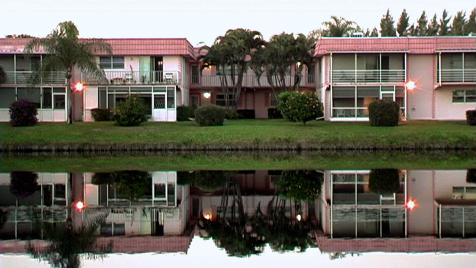Still of the film Kings Point. A full shot of two pink houses next to each other, there are two trees between them. Their reflection is seen in the body of water in front of them.