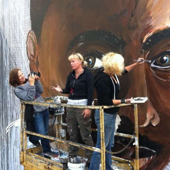 Three women standing on a scaffold in front of a large painting of a man's face, with one woman filming another speaking while the last one paints