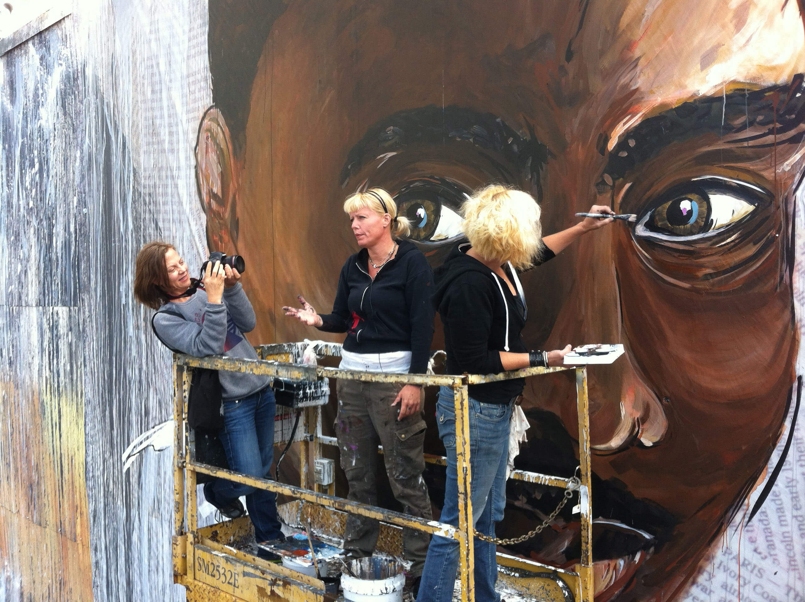 Three women standing on a scaffold in front of a large painting of a man's face, with one woman filming another speaking while the last one paints