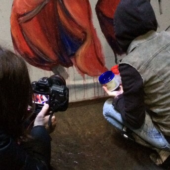 The backs of two women crouching in front of a painting with orange and blue, with one woman filming the other painting.
