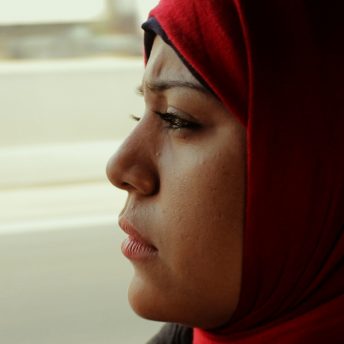 Still from The Trials of Spring. A person in a headscarf looking out the window of a moving vehicle. Color photograph.