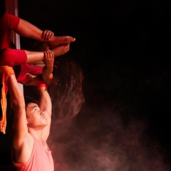 Still from Even When I Fall. A man is supporting a woman as she hangs sideways in the air suspended by canopy silks. The woman's arms are stretched backwards as she holds onto her ankles and her face is hidden behind her hair. The two figures are illuminated against a black background.