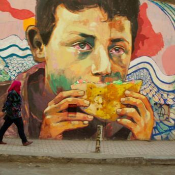 Still from The Trials of Spring. A large colorful mural of a person holding a pita with red eyes. On the sidewalk, a person is walking and looking up at the mural. Color photograph.