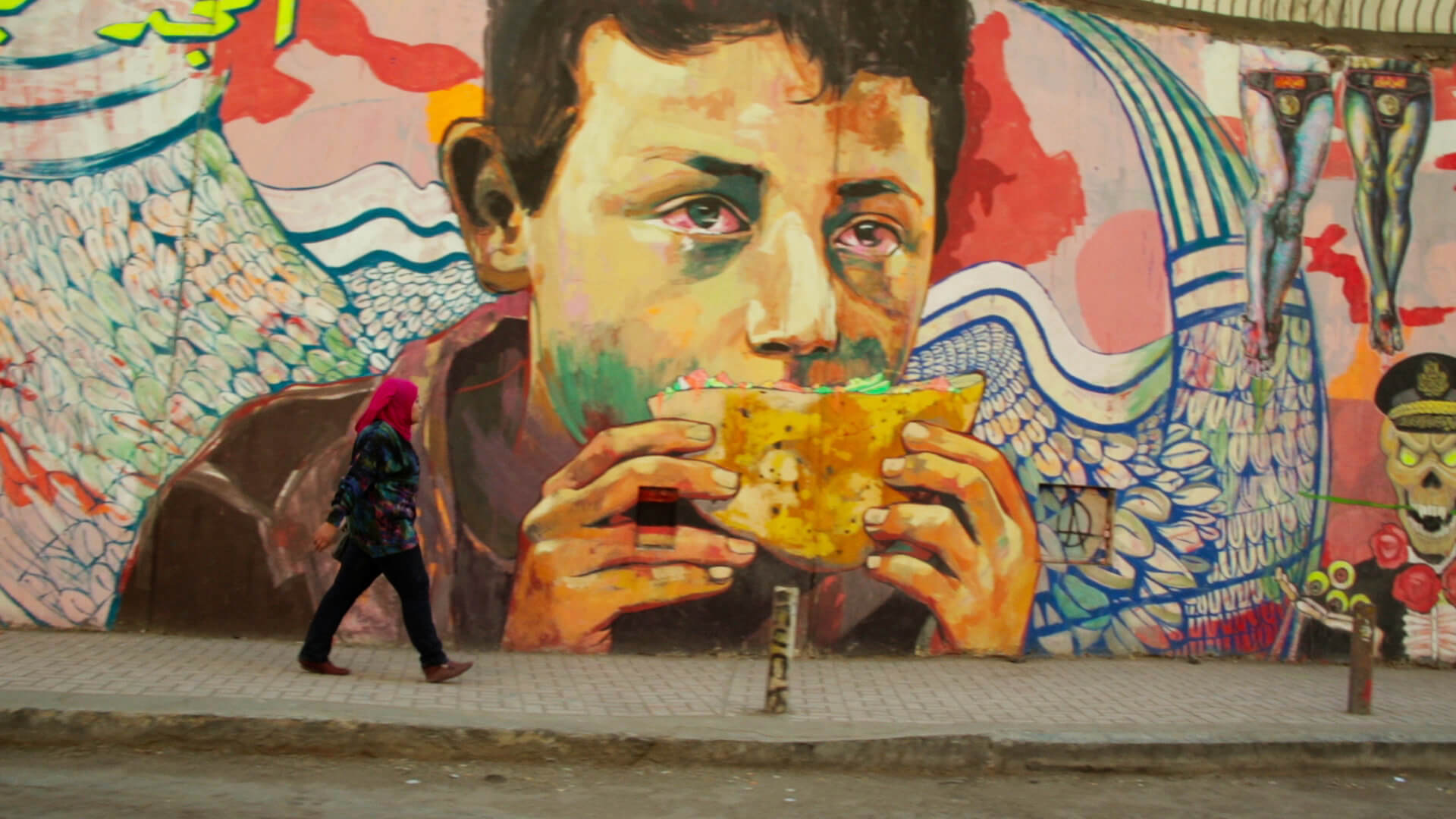 Still from The Trials of Spring. A large colorful mural of a person holding a pita with red eyes. On the sidewalk, a person is walking and looking up at the mural. Color photograph.