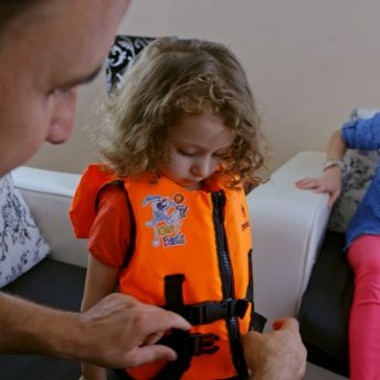 Still from It Will Be Chaos. A child is wearing a bright orange life vest and is looking down as a man is buckling the straps of the vest. A young girl is looking on.