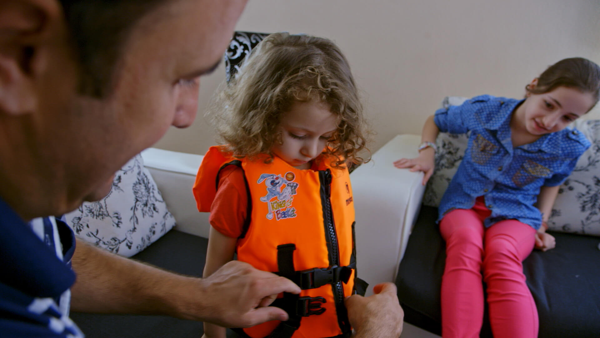 Still from It Will Be Chaos. A child is wearing a bright orange life vest and is looking down as a man is buckling the straps of the vest. A young girl is looking on.