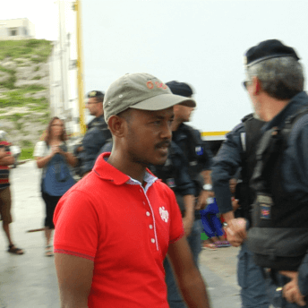 Still from It Will Be Chaos. A man wearing a bright red polo shirt and baseball hat is walking. In the background, there are people who are not in focus. There is a man filming with a large camera on his shoulder and there are several men dressed in military clothes, wearing black utility vests and black berets.