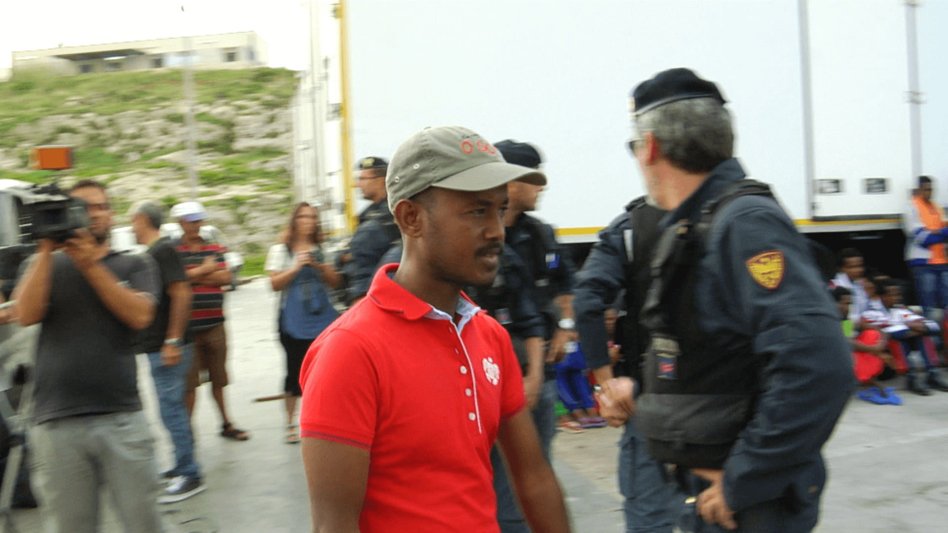 Still from It Will Be Chaos. A man wearing a bright red polo shirt and baseball hat is walking. In the background, there are people who are not in focus. There is a man filming with a large camera on his shoulder and there are several men dressed in military clothes, wearing black utility vests and black berets.