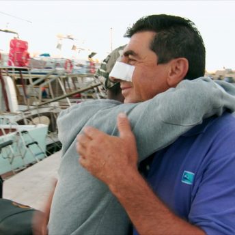 Still from It Will Be Chaos. Two people are hugging. They are seen from the side, and one man has a bandage on his nose and is smiling. The other person's face is not visible. A line of docked boats is seen behind them.