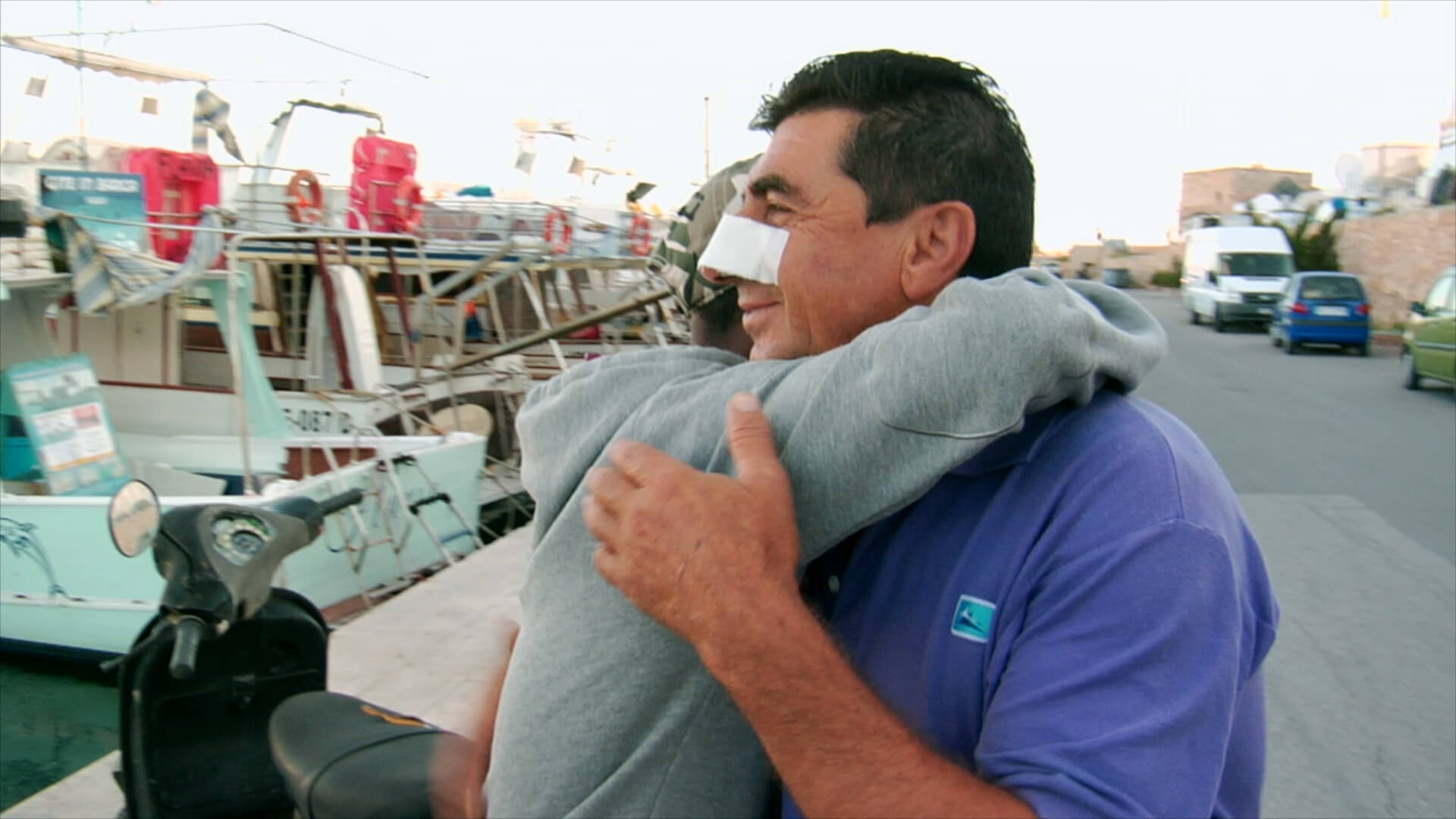 Still from It Will Be Chaos. Two people are hugging. They are seen from the side, and one man has a bandage on his nose and is smiling. The other person's face is not visible. A line of docked boats is seen behind them.