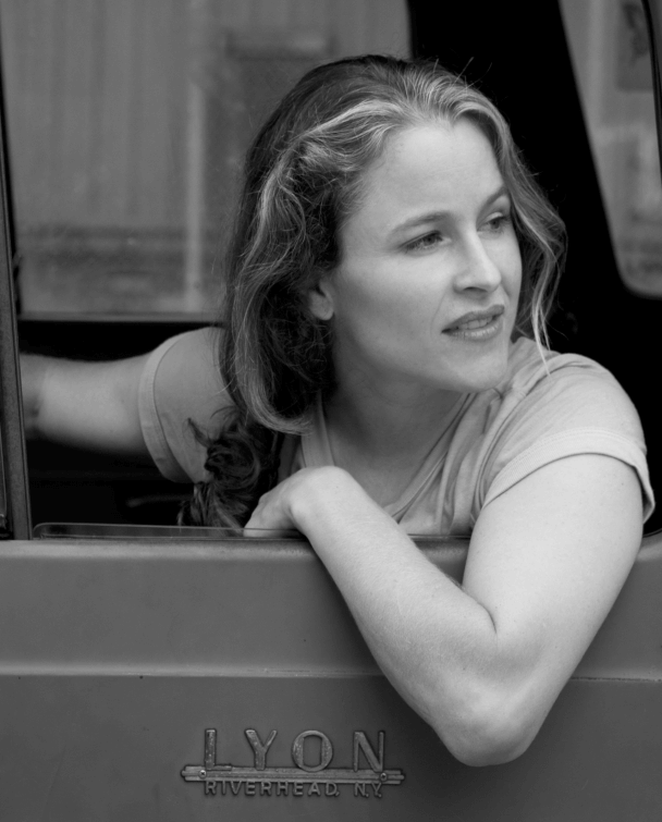 Jesse Epstein has curly hair and is leaning out the window of a car and looking to her right. Portrait in black and white.