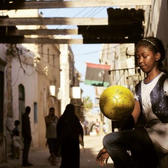 Still from Freedom Fields. A girl, illuminated by the sun, is bouncing a yellow soccer ball on her knee. She is wearing jeans, a white t-shirt, and a sparkly headband and vest. In the background, other people are walking in the street; some looking in the direction of the girl. The Libyan flag is illuminated above a doorway.