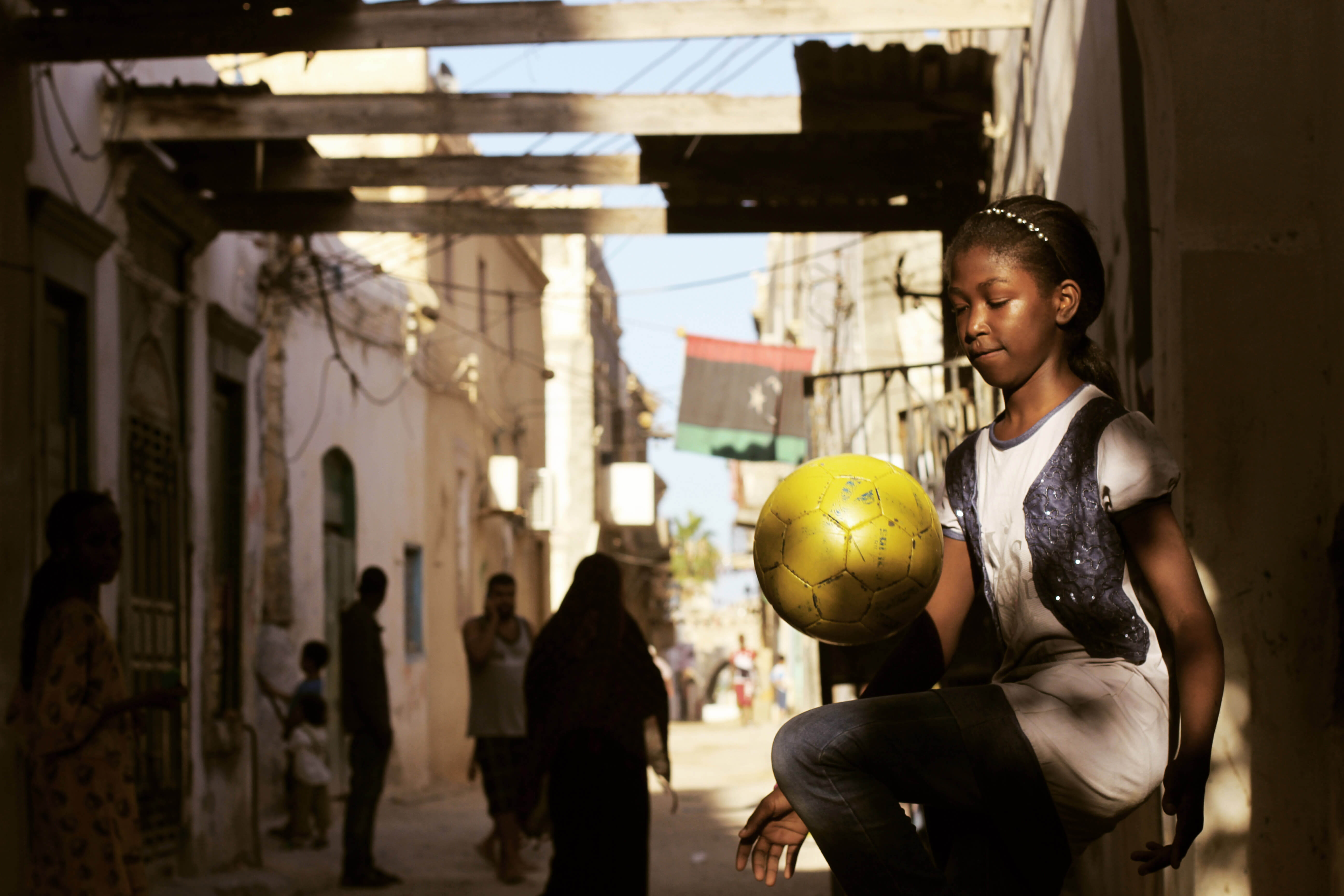 Still from Freedom Fields. A girl, illuminated by the sun, is bouncing a yellow soccer ball on her knee. She is wearing jeans, a white t-shirt, and a sparkly headband and vest. In the background, other people are walking in the street; some looking in the direction of the girl. The Libyan flag is illuminated above a doorway.