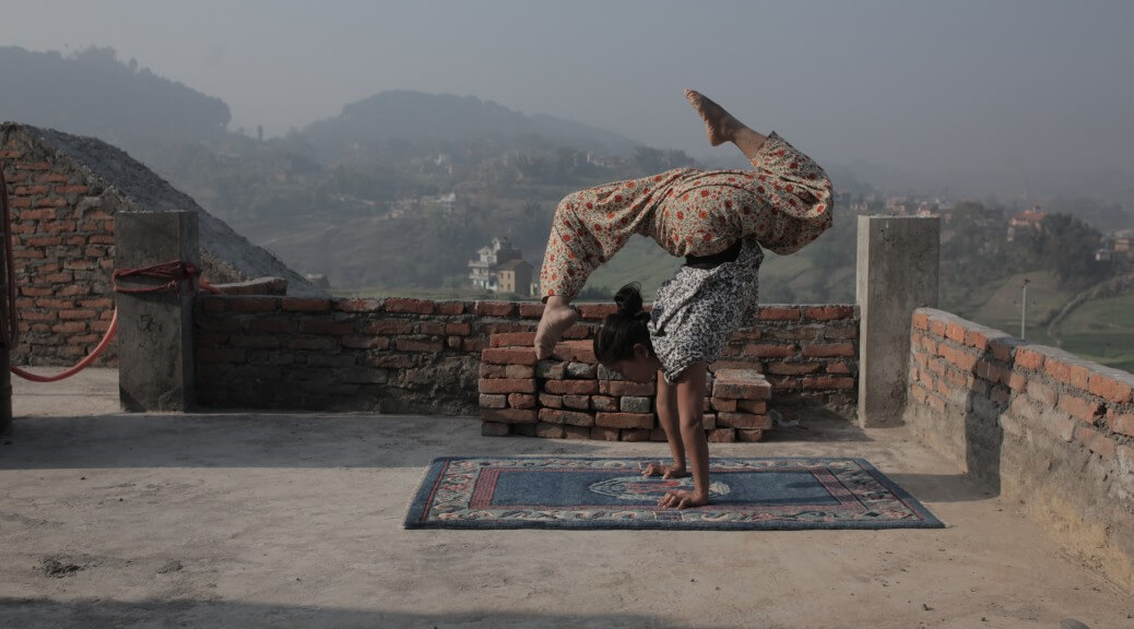 Still from Even When I Fall. A young woman is upside down on a blue Persian-style rug. She is balancing on her arms, with one leg lifted in front of her head, and the other leg in the air. She is wearing a black-and white patterned shirt and brightly-partterned pants. She is on a rooftop; houses, fields, and forested hills are seen in the distance behind her.