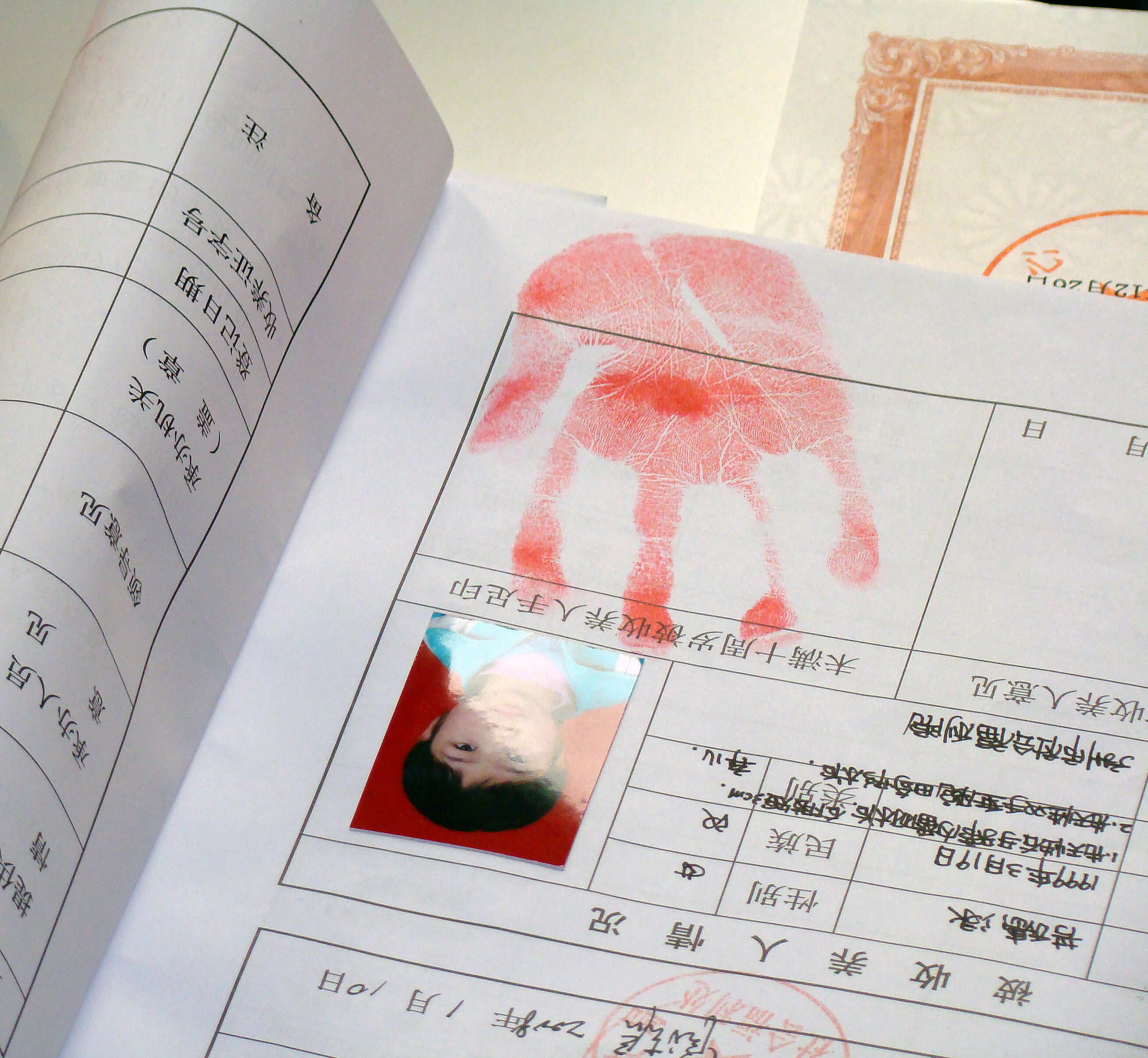 An upside-down Chinese document with a portrait of a young kid affixed to it. A red handprint is on the page. Color photograph.