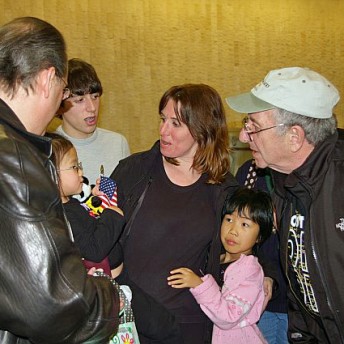 A group of people stands close to each other, with one child holding onto a woman wearing all black, and another child holding a small American flag.