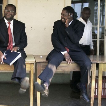 Two men in business attire sitting on a table. One of them holds some papers and the other one is talking via cellphone.