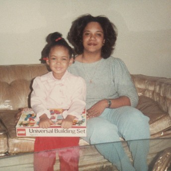 A woman and a young girl are sitting on a sofa and both smile at the camera. The girl has a box with a game on her lap.