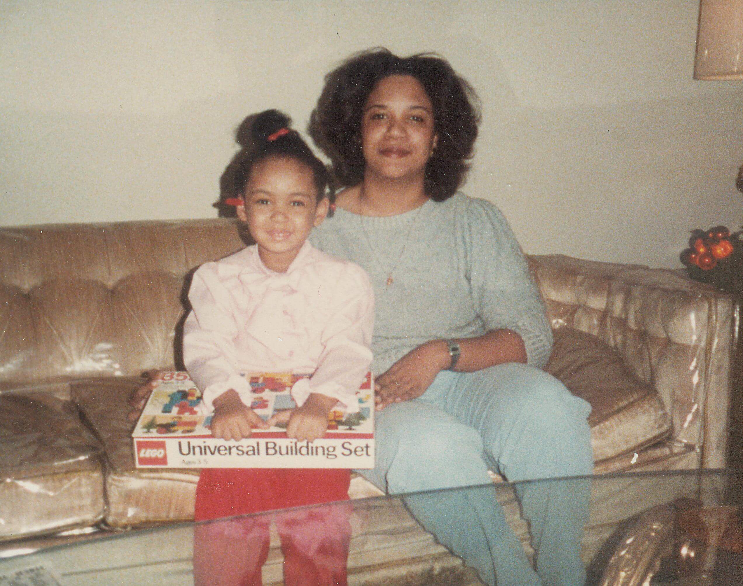 A woman and a young girl are sitting on a sofa and both smile at the camera. The girl has a box with a game on her lap.