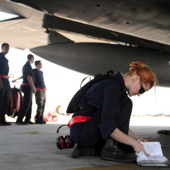 Still from The Invisible War. A person crouches on the ground in front of a large plane in the background. They are looking at a binder full of documents. They have a sleek bun and are wearing dark sunglasses. A pair of noise-block headphones rest on the ground behind them, and three people stand off to the side in the distance.
