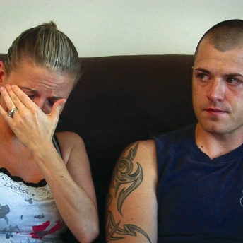 Still from The Invisible War. Two people sit on a brown couch shoulder to shoulder, facing the camera. The person on the left has their head in their hands, blocking their face from view. The person on the right looks over at the person on the left.
