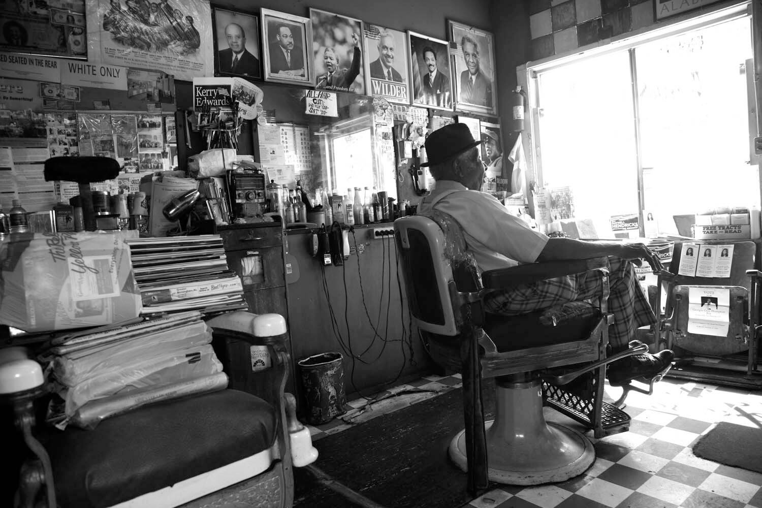 A still from The Barber of Birmingham. A man sits in a barbers chair, turned away from the camera. The wall behind him is covered in headshots, and various ephemera are stacked on the surfaces around him. Photo in black and white.