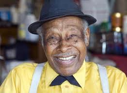 A close-up photo of a man with a big smile on his face. He wears a yellow shirt, white suspenders, a black bowtie, and a black fedora.