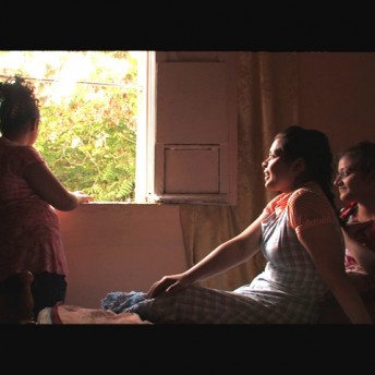 Still from Between Earth & Sky. A woman is standing looking outside a window, while two other women are sitting on a bed and looking at her.