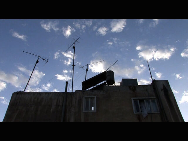 Still from Between Earth & Sky. Low angle shot of three consecutive apartments with antennas on their rooftops.