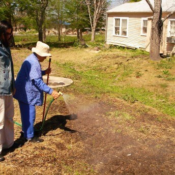 Still from My Louisiana Love. A young woman stands beside an older woman that wears a hat, watching her water a patch of dirt on a lawn.