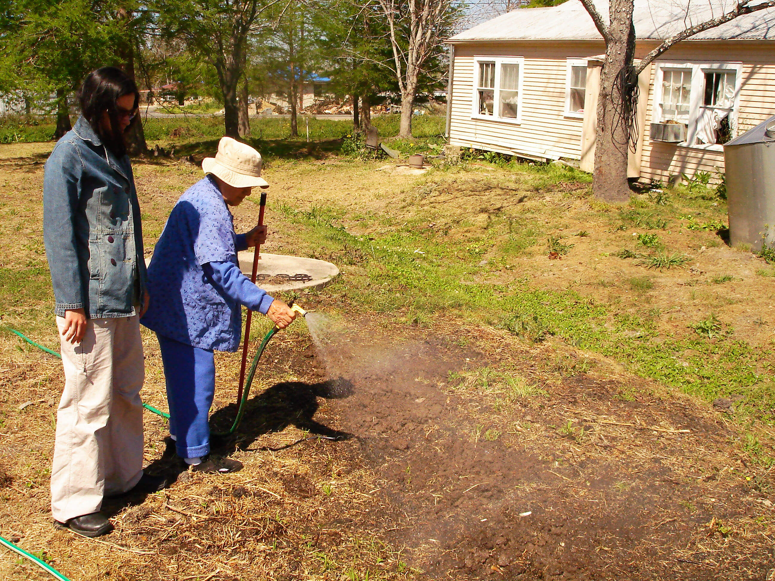 Still from My Louisiana Love. A young woman stands beside an older woman that wears a hat, watching her water a patch of dirt on a lawn.