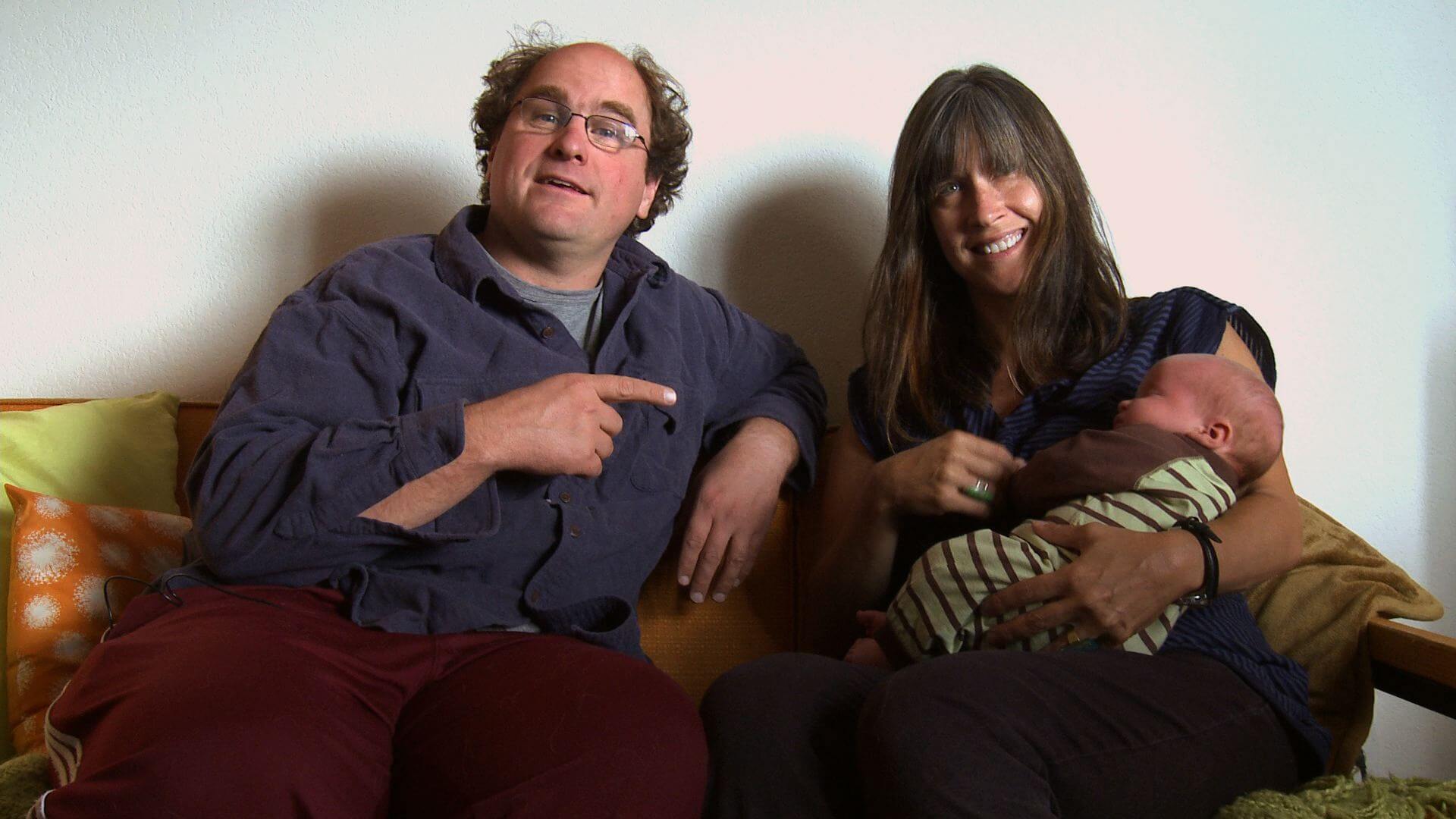 Still from Bag It. A couple is sitting on a couch. The man points at the woman. The woman is feeding a baby in her arms.