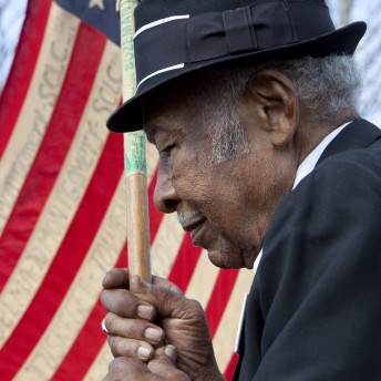A close-up photo of a man in a black fedora holding an American flag. He has short grey hair.