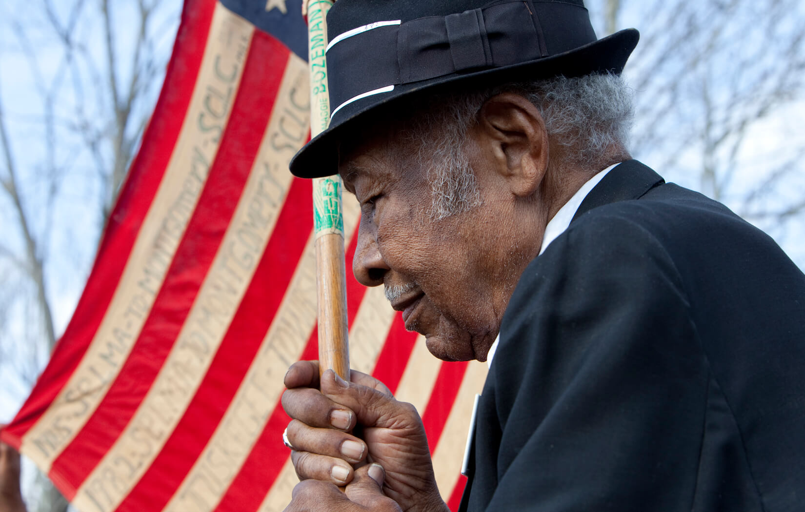 A close-up photo of a man in a black fedora holding an American flag. He has short grey hair.