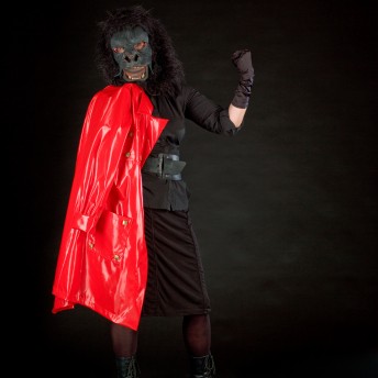 A person wears a gorilla mask, and an all-black outfit, and holds a red latex coat. The background is black.