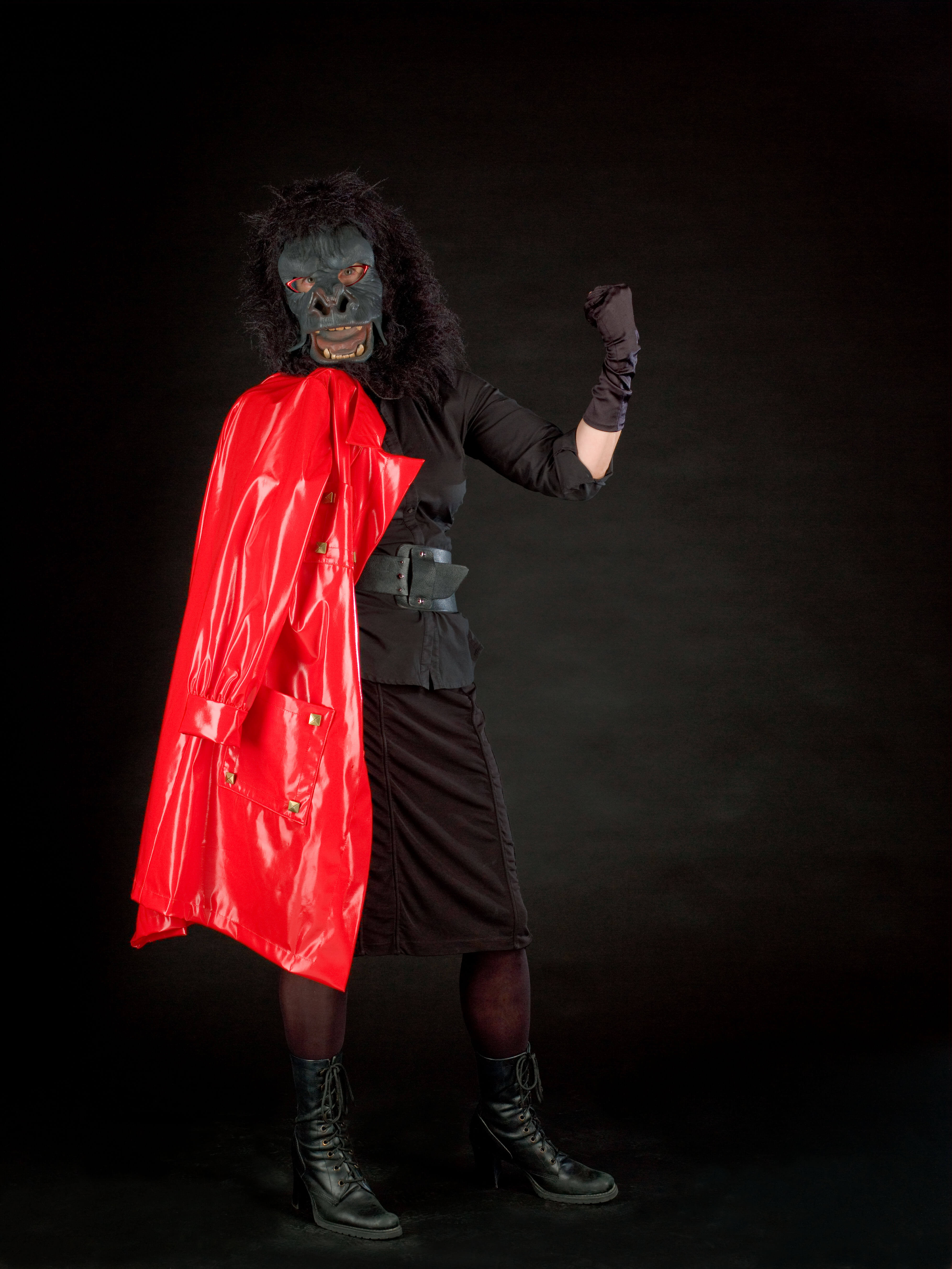 A person wears a gorilla mask, and an all-black outfit, and holds a red latex coat. The background is black.
