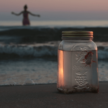 Still from Girls of the Moon. In the foreground, close up of a glass jar filled with water, pebbles, and a gold fish, sits on the sand. Part of the light in the jar refracts the orange light of the sunset. In the out of focus background, waves are rolling on the shore, and a woman is standing in the water, with her arms outstretched.