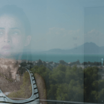 Still from Girls of the Moon. A young woman is staring through a window. She has dark hair, pulled back into a ponytail. She is wearing pink lipstick, a black and white striped tank top, and a gold necklace. She is heavily freckled. In the reflection of the glass is seen a city, water, and mountains.