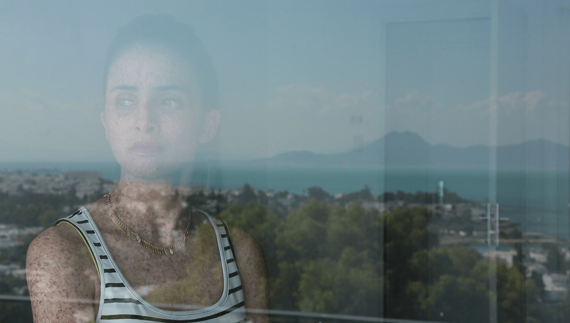 Still from Girls of the Moon. A young woman is staring through a window. She has dark hair, pulled back into a ponytail. She is wearing pink lipstick, a black and white striped tank top, and a gold necklace. She is heavily freckled. In the reflection of the glass is seen a city, water, and mountains.