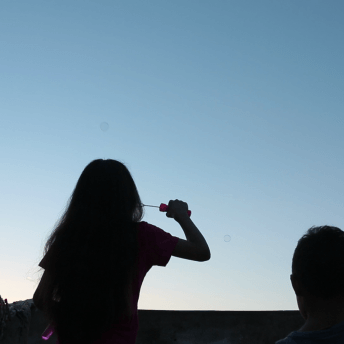 Still from Girls of the Moon. Silhouette of two children -- a girl and a boy -- blowing bubbles. They are seen from behind, the sky is light blue and goes to yellow closer to the horizon, as if it is sunrise or sunset.