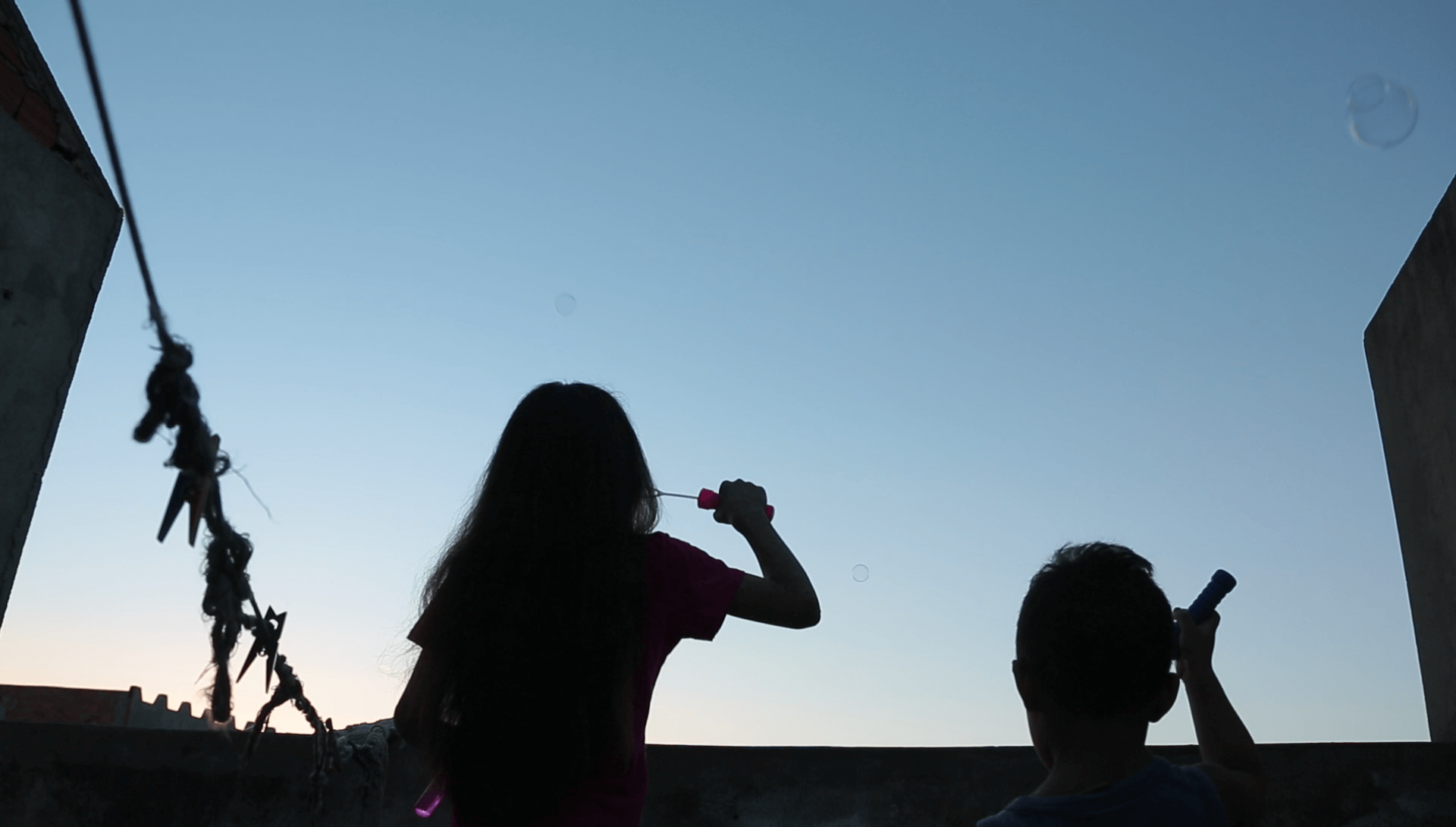 Still from Girls of the Moon. Silhouette of two children -- a girl and a boy -- blowing bubbles. They are seen from behind, the sky is light blue and goes to yellow closer to the horizon, as if it is sunrise or sunset.