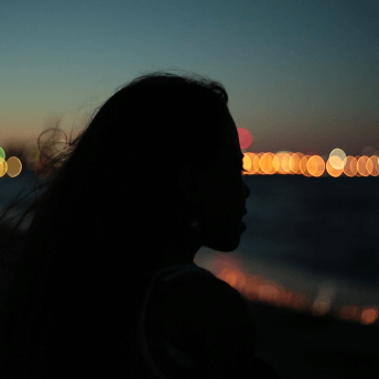 Still from Girls of the Moon. Backlighted silhouette of a woman with long hair in front of a waterfront. On the horizon, there is a string of out of focus lights, which are reflected in the water.